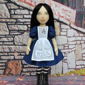 May include: A doll dressed in a blue and white dress with a white apron. The doll has long black hair and is wearing black boots. The doll is standing in front of a stone wall with a sign that says "Entrance".