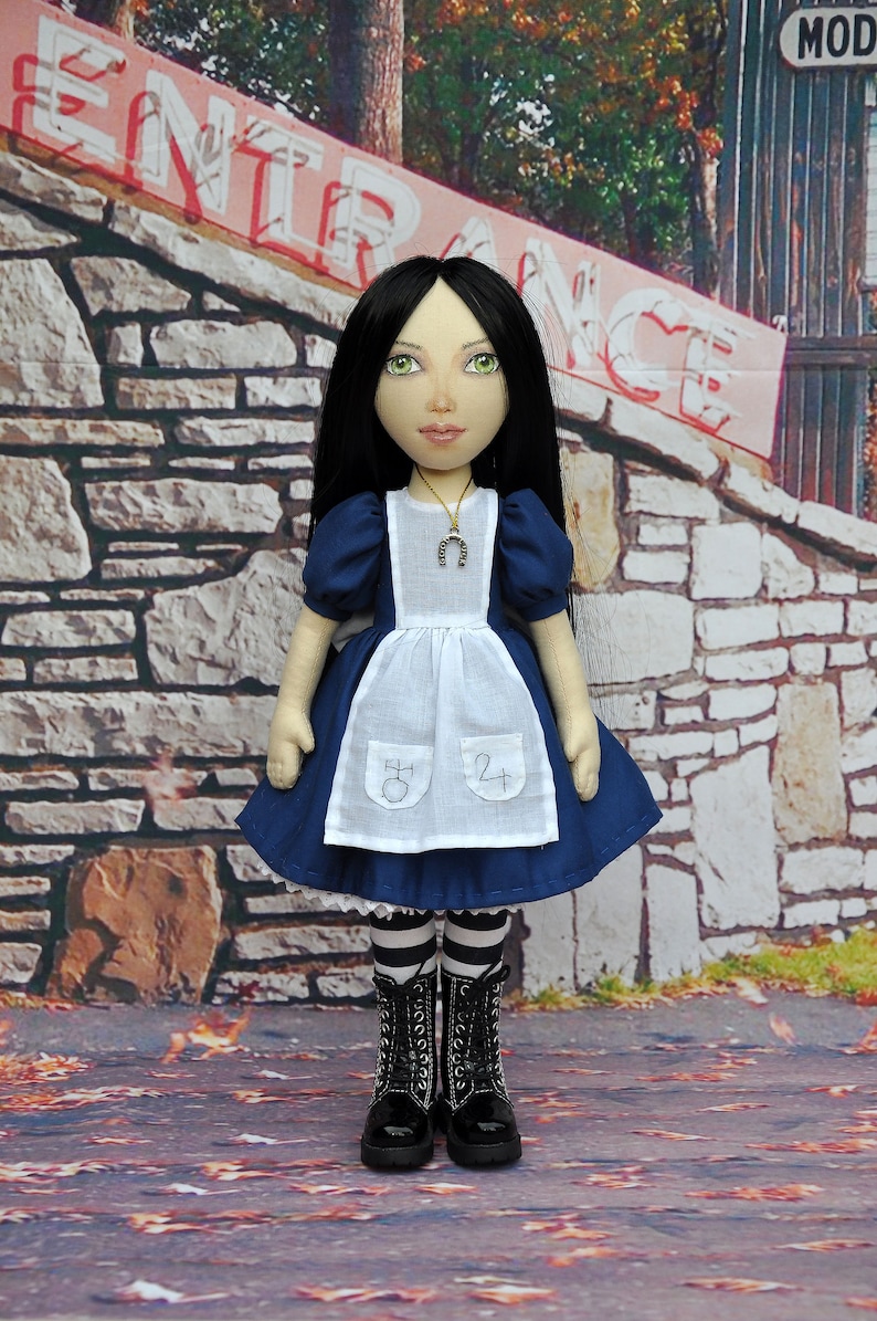 May include: A doll dressed in a blue and white dress with a white apron. The doll has long black hair and is wearing black boots. The doll is standing in front of a stone wall with a sign that says "Entrance".