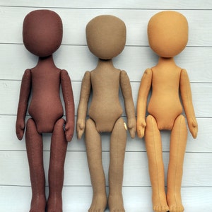 May include: Three fabric dolls standing in a row. The doll on the left is brown, the middle doll is tan, and the doll on the right is light brown. All three dolls have no facial features and are standing on a white wooden surface.