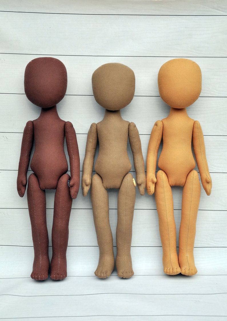 May include: Three fabric dolls standing in a row. The doll on the left is brown, the middle doll is tan, and the doll on the right is light brown. All three dolls have no facial features and are standing on a white wooden surface.