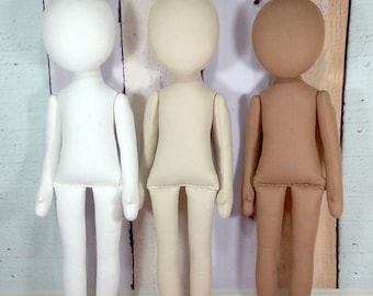 Op de afbeelding: Drie stoffen poppen in wit, beige en bruin. De poppen hebben ronde hoofden, eenvoudige lichamen en beweegbare armen en benen. Ze staan rechtop tegen een lichte achtergrond van houten panelen, wat hun potentieel voor personalisatie aantoont.