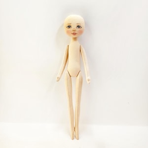 Puede incluir: Una muñeca de tela blanca lisa con la cara pintada. La muñeca tiene ojos marrones, mejillas rosadas y boca roja. La muñeca está de pie sobre un fondo blanco.