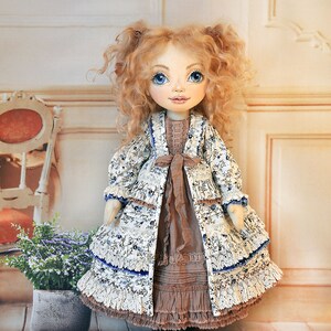 May include: A handmade doll with curly blonde hair, blue eyes, and a white dress with blue floral print and brown accents. The doll is wearing a white jacket with blue floral print and a brown sash. The doll is standing on a wooden floor.