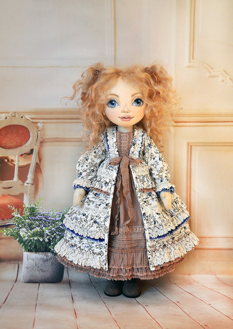 May include: A handmade doll with curly blonde hair, blue eyes, and a white dress with blue floral print and brown accents. The doll is wearing a white jacket with blue floral print and a brown sash. The doll is standing on a wooden floor.