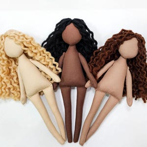 May include: Three handmade cloth dolls with different skin tones and hair colors. The dolls have beige bodies, with one doll having blonde curly hair, one with black curly hair, and one with brown curly hair. The dolls are standing upright.