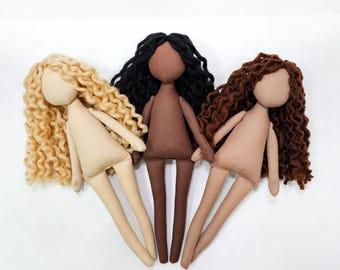 May include: Three handmade cloth dolls with different skin tones and hair colors. The dolls have beige bodies, with one doll having blonde curly hair, one with black curly hair, and one with brown curly hair. The dolls are standing upright.