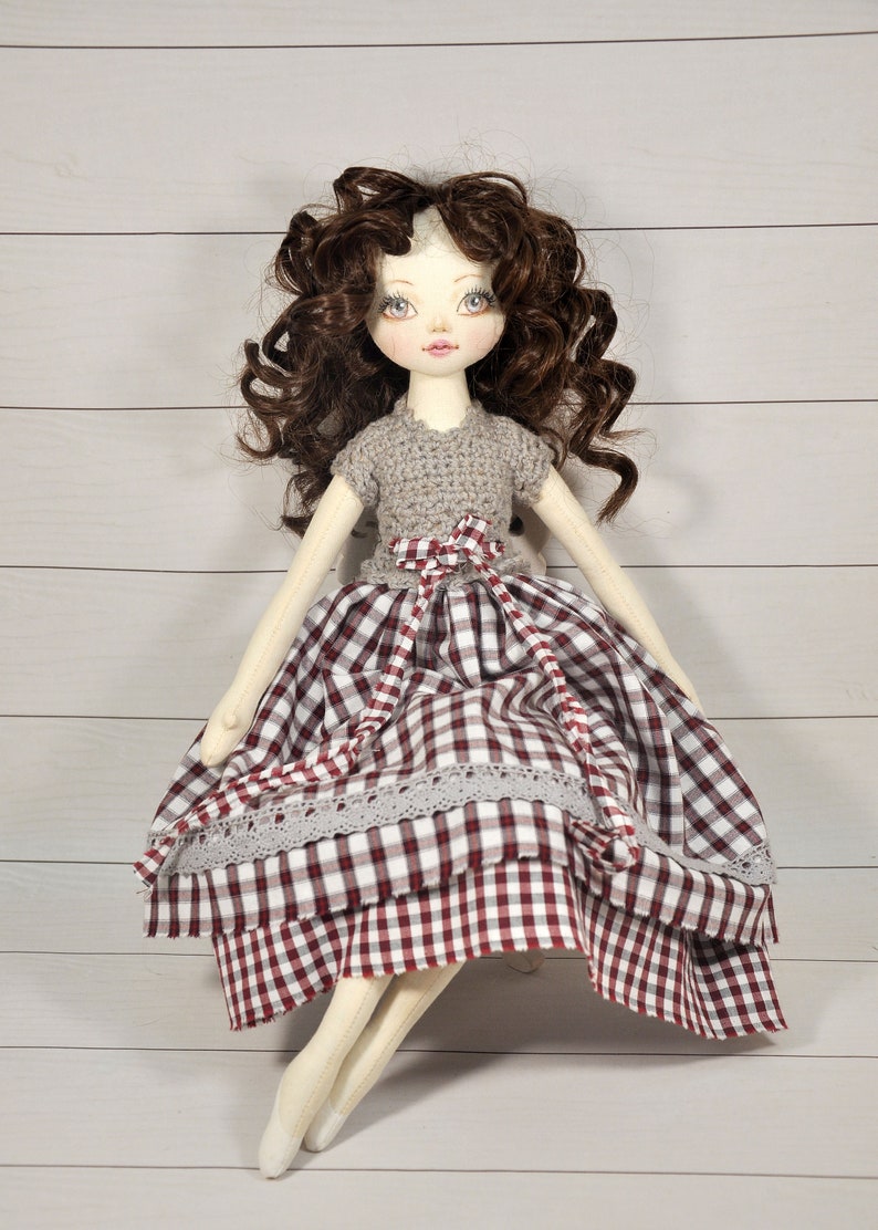 Puede incluir: Una mu&ntilde;eca hecha a mano con cabello casta&ntilde;o rizado, que lleva un top de crochet gris y una falda de cuadros rojos y blancos con ribete de encaje.