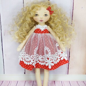May include: A handmade doll with blonde curly hair, wearing a red dress with white polka dots and white lace trim. The doll has a red bow in her hair and is standing on a pink wooden floor.