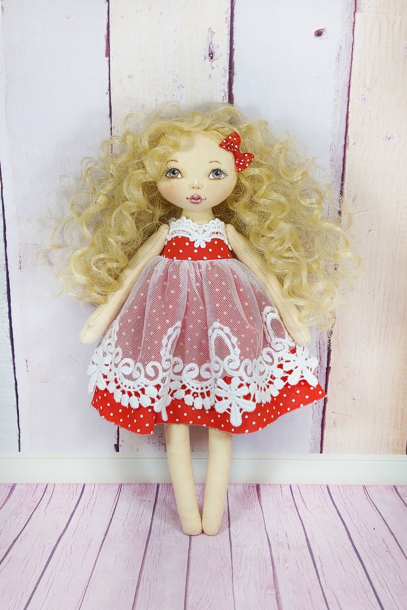 May include: A handmade doll with blonde curly hair, wearing a red dress with white polka dots and white lace trim. The doll has a red bow in her hair and is standing on a pink wooden floor.