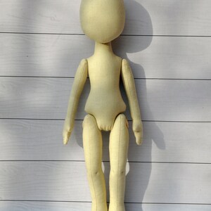 May include: A plain, unpainted, beige fabric doll with a round head and no facial features. The doll has long arms and legs, and is standing on a white wooden surface.