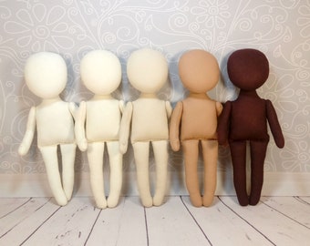 May include: Five fabric doll bodies in varying skin tones. The dolls have simple, rounded heads, arms, and legs. The colors range from cream to dark brown, suggesting a diverse representation. They stand against a light gray floral wallpaper.