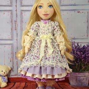 May include: A handmade doll with long blonde hair, wearing a purple and white floral dress with a yellow sash and lace trim. The doll is wearing purple shoes and is standing on a wooden floor.