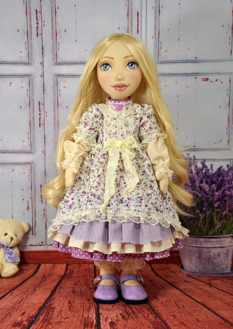 May include: A handmade doll with long blonde hair, wearing a purple and white floral dress with a yellow sash and lace trim. The doll is wearing purple shoes and is standing on a wooden floor.