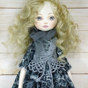 Puede incluir: Una mu&ntilde;eca hecha a mano con cabello rubio rizado, que lleva un chaleco de crochet gris y una bufanda gris sobre un vestido estampado en gris y blanco.