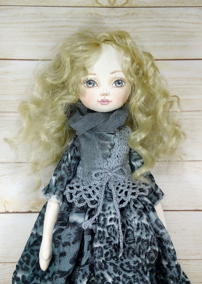 Puede incluir: Una mu&ntilde;eca hecha a mano con cabello rubio rizado, que lleva un chaleco de crochet gris y una bufanda gris sobre un vestido estampado en gris y blanco.