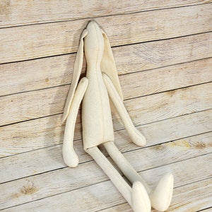 May include: A white fabric bunny rabbit toy with long ears and long limbs sitting on a light brown wooden surface.