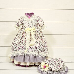 K&ouml;nnte beinhalten: Ein wei&szlig;es Kleid mit lila Blumenmuster und Spitzenbesatz. Das Kleid hat ein gelbes Band, das in der Taille gebunden ist. Ein passender wei&szlig;er Hut mit lila Blumenmuster und Spitzenbesatz ist ebenfalls enthalten.