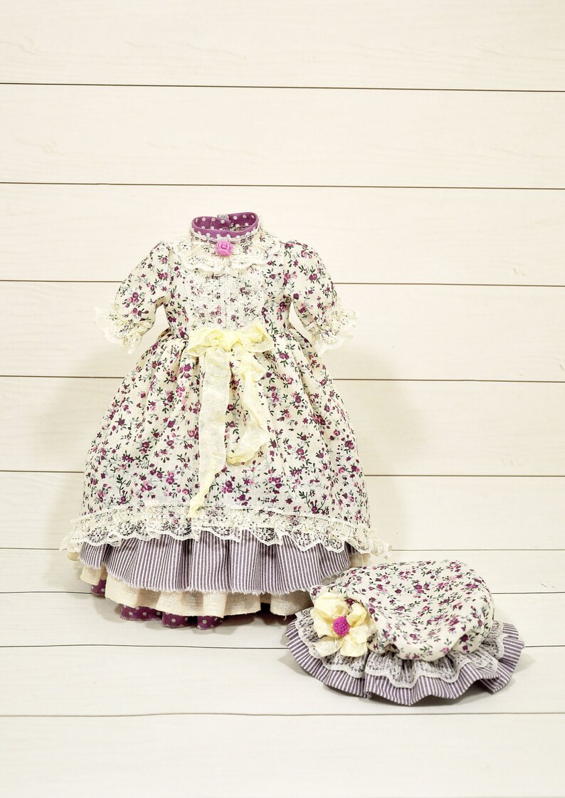 K&ouml;nnte beinhalten: Ein wei&szlig;es Kleid mit lila Blumenmuster und Spitzenbesatz. Das Kleid hat ein gelbes Band, das in der Taille gebunden ist. Ein passender wei&szlig;er Hut mit lila Blumenmuster und Spitzenbesatz ist ebenfalls enthalten.