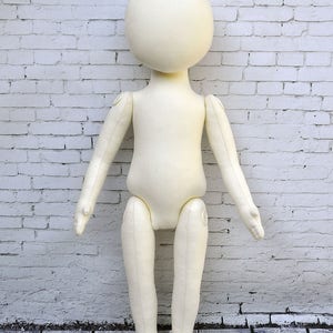 K&ouml;nnte beinhalten: Eine wei&szlig;e Stoffpuppe mit beweglichen Armen und Beinen. Die Puppe steht auf einem Holzboden vor einer wei&szlig;en Backsteinwand.