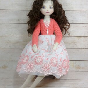 Puede incluir: Una mu&ntilde;eca hecha a mano con el pelo largo y rizado marr&oacute;n, que lleva un vestido blanco con bordados florales rosas y un c&aacute;rdigan de punto coral. La mu&ntilde;eca tiene una cara pintada con ojos azules y labios rosas.