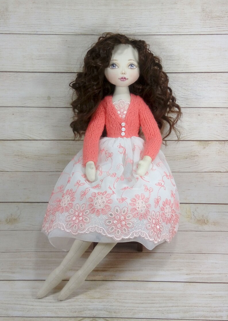 Puede incluir: Una mu&ntilde;eca hecha a mano con el pelo largo y rizado marr&oacute;n, que lleva un vestido blanco con bordados florales rosas y un c&aacute;rdigan de punto coral. La mu&ntilde;eca tiene una cara pintada con ojos azules y labios rosas.