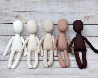 May include: Five fabric dolls in various shades of brown, beige, and white, sitting in a row on a wooden surface. The dolls have no facial features and are made of a soft, plush fabric.
