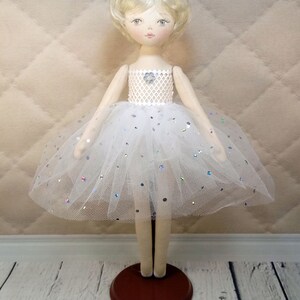 Op de afbeelding: Een witte porseleinen pop met blond haar die een witte tule tutu draagt met zilveren glitter. De pop draagt een kroon en staat op een bruine houten voet.