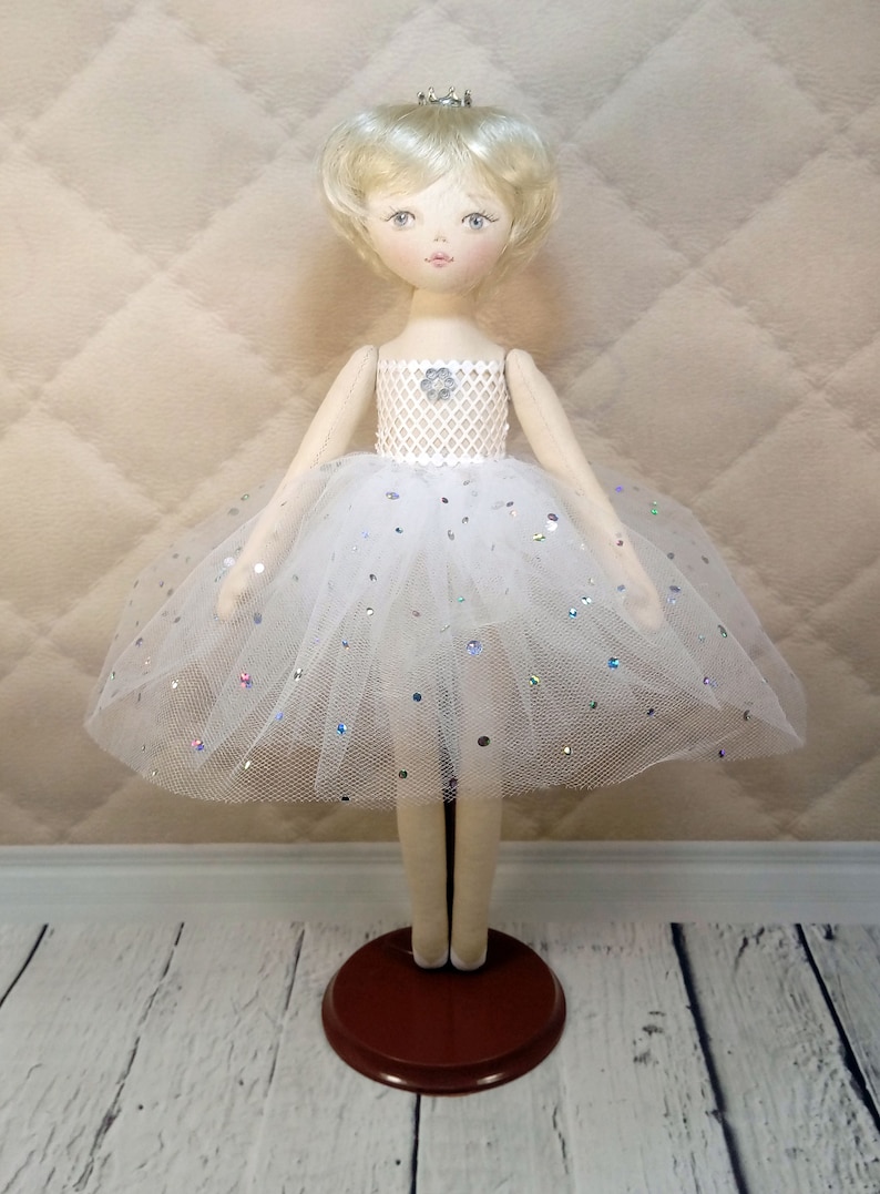 Op de afbeelding: Een witte porseleinen pop met blond haar die een witte tule tutu draagt met zilveren glitter. De pop draagt een kroon en staat op een bruine houten voet.
