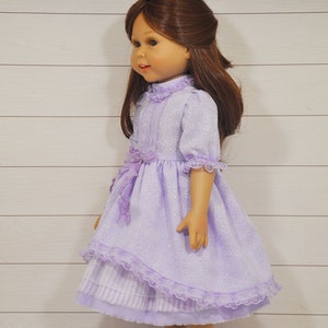 K&ouml;nnte beinhalten: Eine Puppe tr&auml;gt ein lavendelfarbenes Kleid mit wei&szlig;em Spitzenbesatz und passenden Schuhen. Das Kleid hat einen R&uuml;schenkragen und eine Schleife an der Taille.