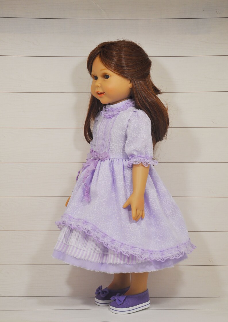 K&ouml;nnte beinhalten: Eine Puppe tr&auml;gt ein lavendelfarbenes Kleid mit wei&szlig;em Spitzenbesatz und passenden Schuhen. Das Kleid hat einen R&uuml;schenkragen und eine Schleife an der Taille.