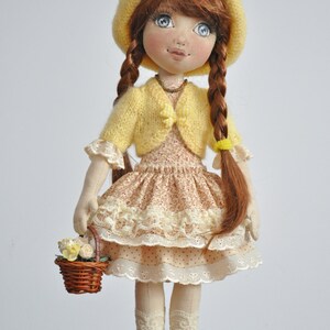 May include: A handmade doll with brown hair, blue eyes, and a yellow knitted hat and cardigan. She is wearing a floral dress with lace trim and white shoes. She is holding a basket of flowers.