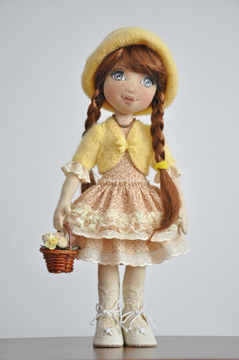 May include: A handmade doll with brown hair, blue eyes, and a yellow knitted hat and cardigan. She is wearing a floral dress with lace trim and white shoes. She is holding a basket of flowers.