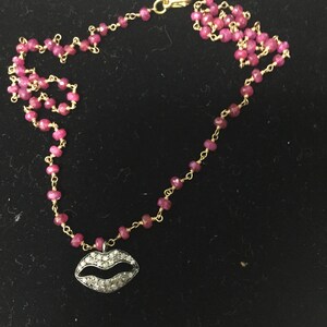 May include: A gold chain necklace with a pink ruby bead design and a diamond-encrusted lip pendant.