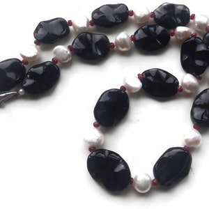 May include: A necklace featuring large, polished black stones, interspersed with white pearls and small red beads. The necklace is strung on a white cord and has a silver clasp. The design is elegant and eye-catching.