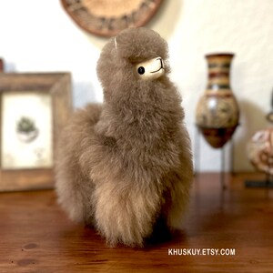 Op de afbeelding: Een bruin alpaca pluche speelgoed met een wit gezicht en zwarte ogen. Het speelgoed staat op een houten oppervlak.