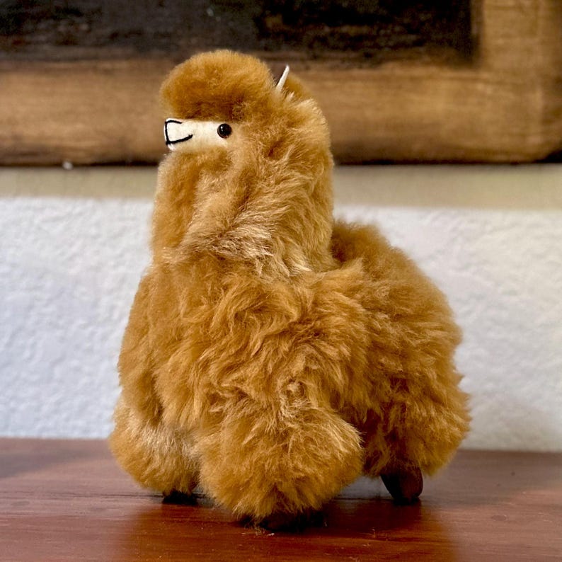 Op de afbeelding: Een bruin alpaca knuffeldier met een pluche textuur. Het speelgoed staat op een houten oppervlak.