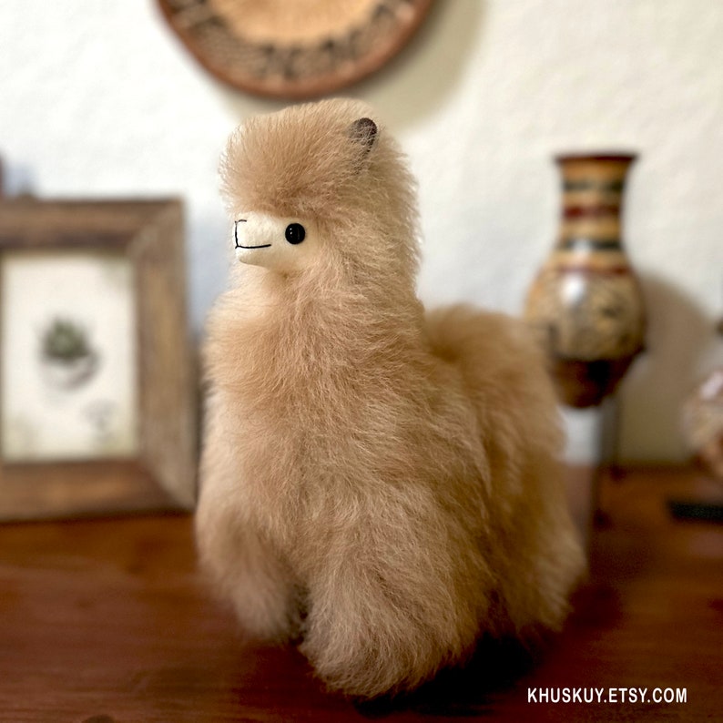 Op de afbeelding: Een lichtbruin alpaca knuffeldier met zwarte ogen en een zwarte neus. Het speelgoed staat op een houten oppervlak.