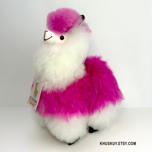 Op de afbeelding: Een roze en wit alpaca knuffeldier met een label met de tekst "De Alpaca by Khuskuy".