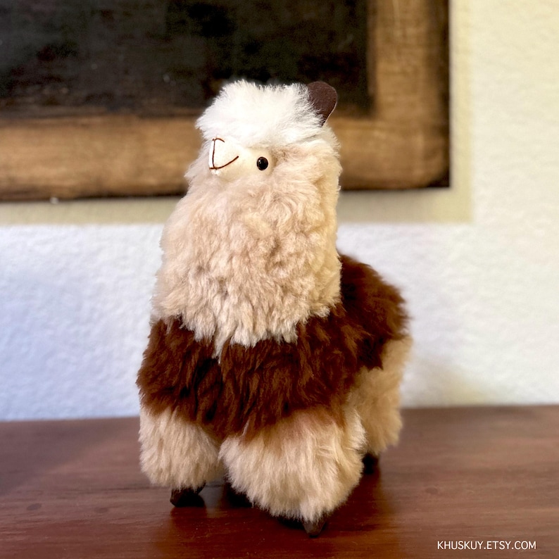Op de afbeelding: Een bruin en wit alpaca pluche speelgoed met een wit gezicht en bruine ogen. Het speelgoed staat op een houten oppervlak.
