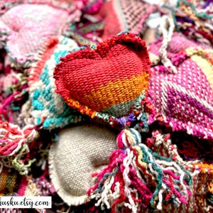 Pode incluir: Close-up de uma coleção de enfeites em forma de coração feitos à mão em várias cores. Os corações são feitos de tecido tecido e apresentam uma variedade de cores, incluindo vermelho, laranja, rosa e verde-azulado. Alguns corações têm borlas. O texto "khusku.etsy.com" é visível.