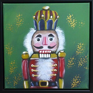 May include: A framed painting of a nutcracker soldier. The nutcracker is dressed in a red coat with gold accents and a tall crown. The background is green with gold leaf-like designs.