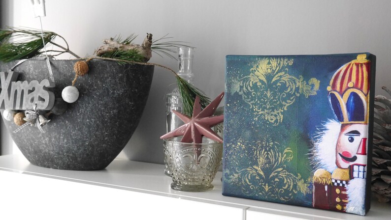 K&ouml;nnte beinhalten: Eine weihnachtliche Dekoration mit einem Leinwandbild eines Nussknackers auf blau-goldenem Hintergrund. Links ein dekorativer Metalltopf mit dem Wort "Xmas". Ein rosa Stern und ein Glaskerzenhalter sind ebenfalls vorhanden.