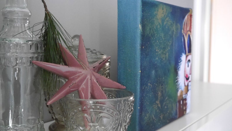 K&ouml;nnte beinhalten: Eine dekorative Anordnung mit einem rosa Sternornament, Glasflaschen und einem kleinen Gem&auml;lde eines Nussknackers. Der Stern befindet sich in einer dekorativen Glasschale. Ein Zweig Gr&uuml;n f&uuml;gt der Pr&auml;sentation eine nat&uuml;rliche Note hinzu.