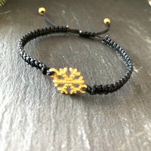 May include: A black braided bracelet with a gold snowflake charm. The bracelet is adjustable with two gold beads at the ends.