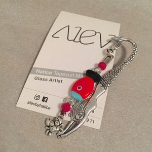 May include: Silver mermaid bookmark with a red and blue fish charm, pink beads, and a silver crab charm. The bookmark is on a white card with the text "Hatice Tepeyurt Mas Glass Artist" and social media handles.
