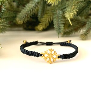 May include: A black adjustable bracelet with a gold snowflake charm. The bracelet is made of cord and has a simple design.