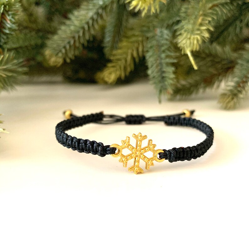 May include: A black adjustable bracelet with a gold snowflake charm. The bracelet is made of cord and has a simple design.