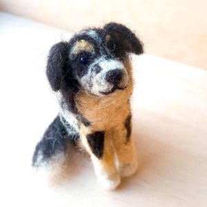 Peut inclure: Une figurine de chien en laine feutrée à l'aiguille avec un pelage tricolore noir, beige et blanc. La petite sculpture représente un chien assis aux oreilles tombantes, aux yeux sombres et à l'aspect texturé et duveteux en fibres de laine.