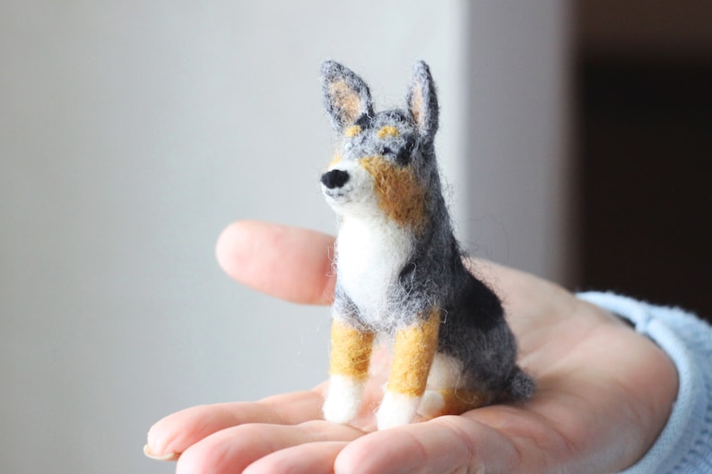 May include: A small, hand-felted dog figurine with black, gray, and tan wool. The dog is sitting upright with its head tilted up.