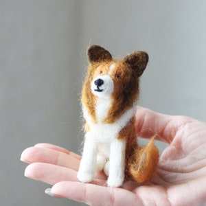 May include: A small, brown and white felt dog figurine with black eyes and a brown nose. The dog is sitting on a person's hand.